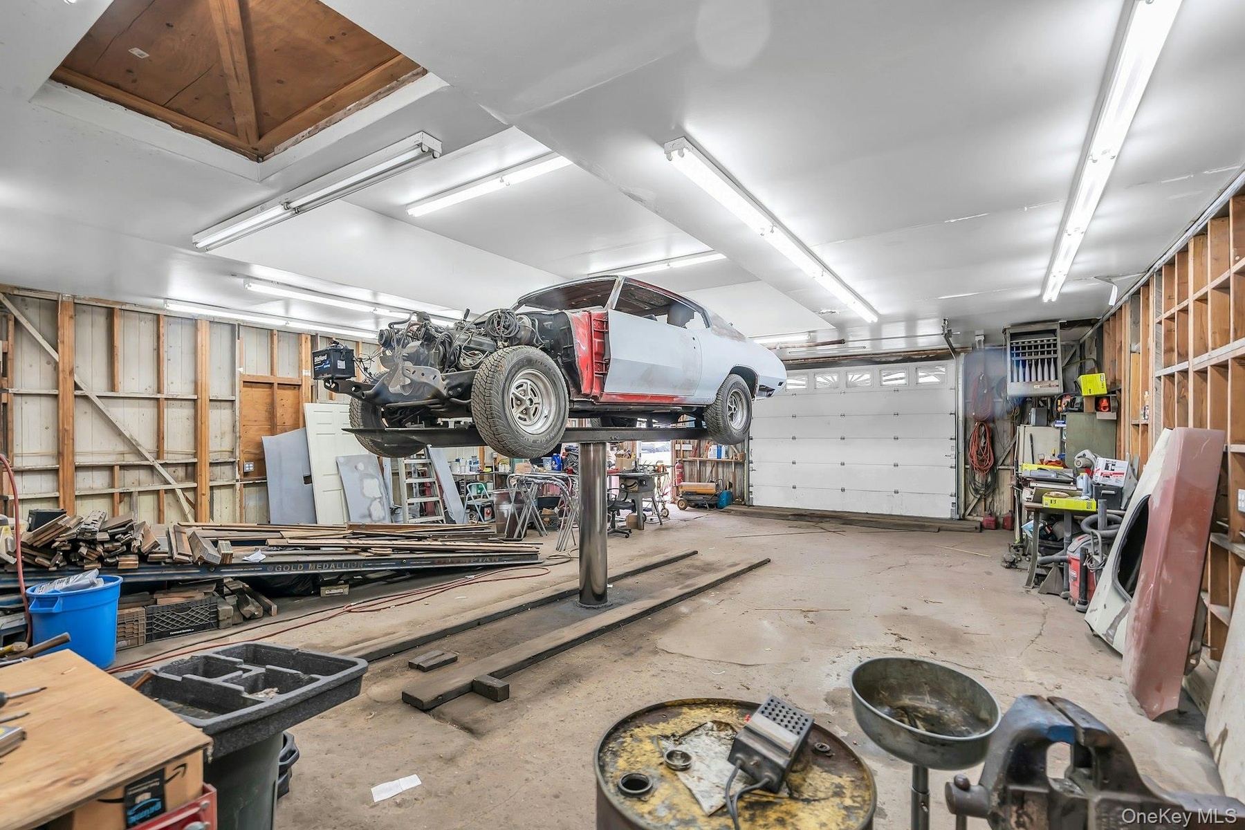 Spacious workshop featuring a built-in automotive lift, overhead fluorescent lighting, exposed wood-stud wall construction, and a multi-panel overhead