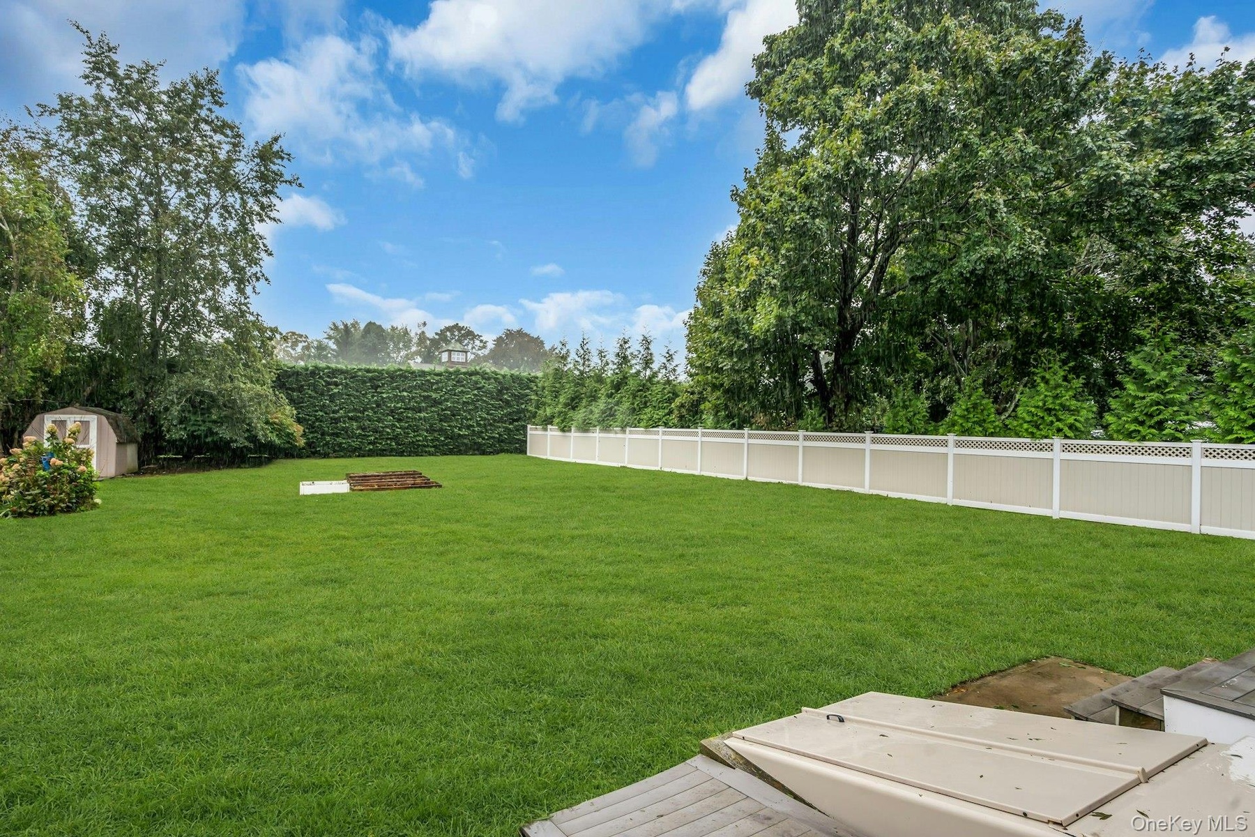 Expansive backyard featuring a level lawn, white vinyl fencing, mature perimeter trees, and a detached storage structure
