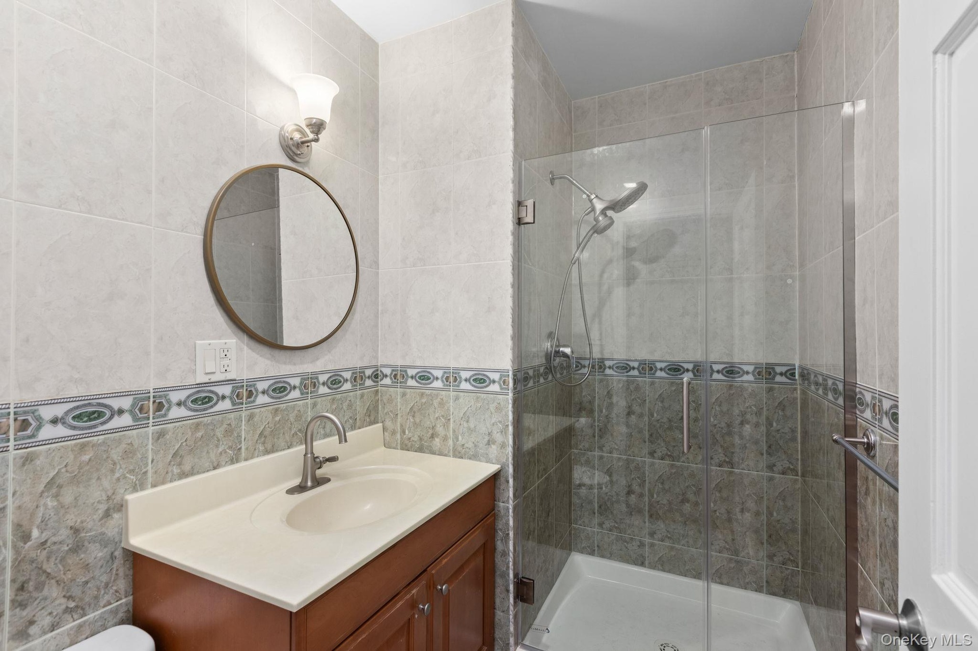 Wood-finish vanity with a molded countertop, glass-enclosed shower featuring a rainfall showerhead, and decorative tiled walls
