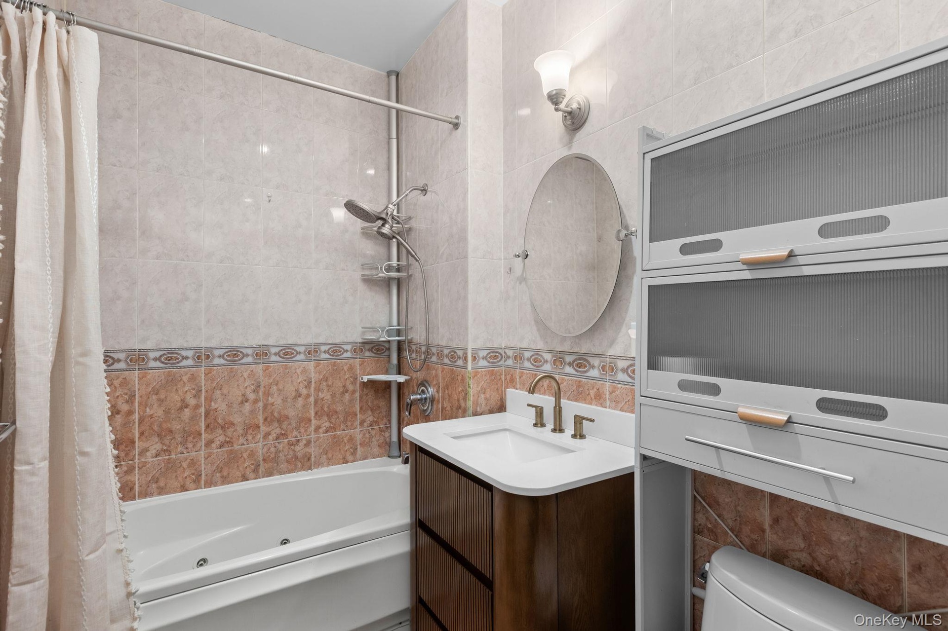 Full-wall tile surround, jetted soaking tub, dark wood-finish vanity with integrated sink, wall-mounted oval mirror, and fixed overhead cabinetry