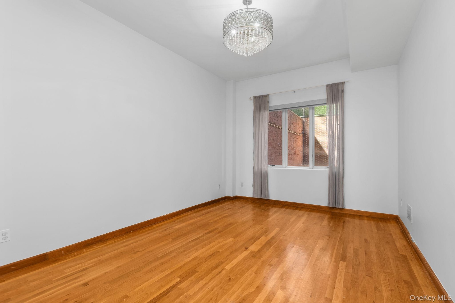 Primary Bedroom featuring wood-finish flooring, a multi-tiered crystal drum chandelier, and a window with exterior brick exposure and blackout shades.