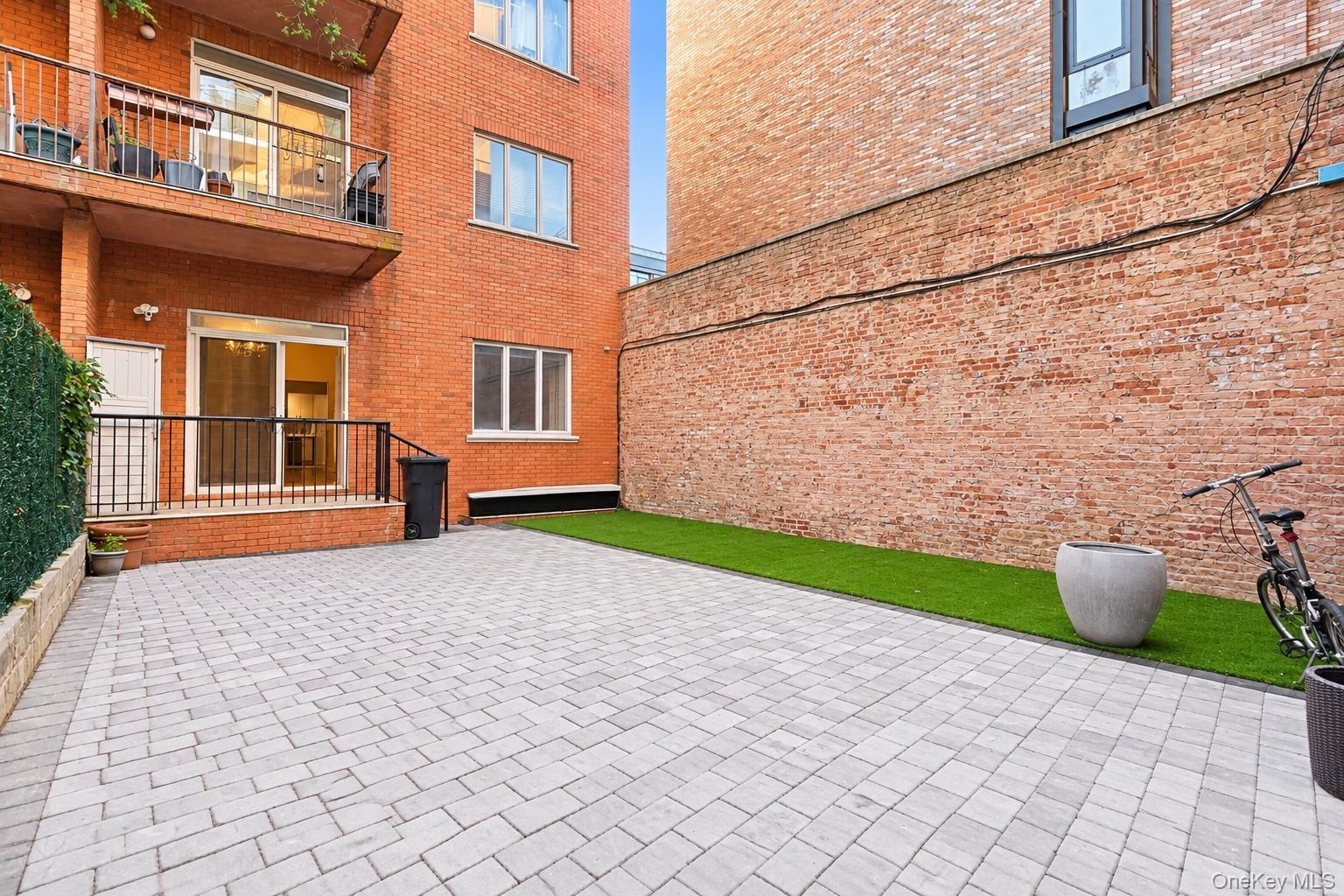 Private outdoor space featuring gray paver hardscaping, a synthetic turf lawn, red brick building exterior, and an elevated metal-railed balcony.