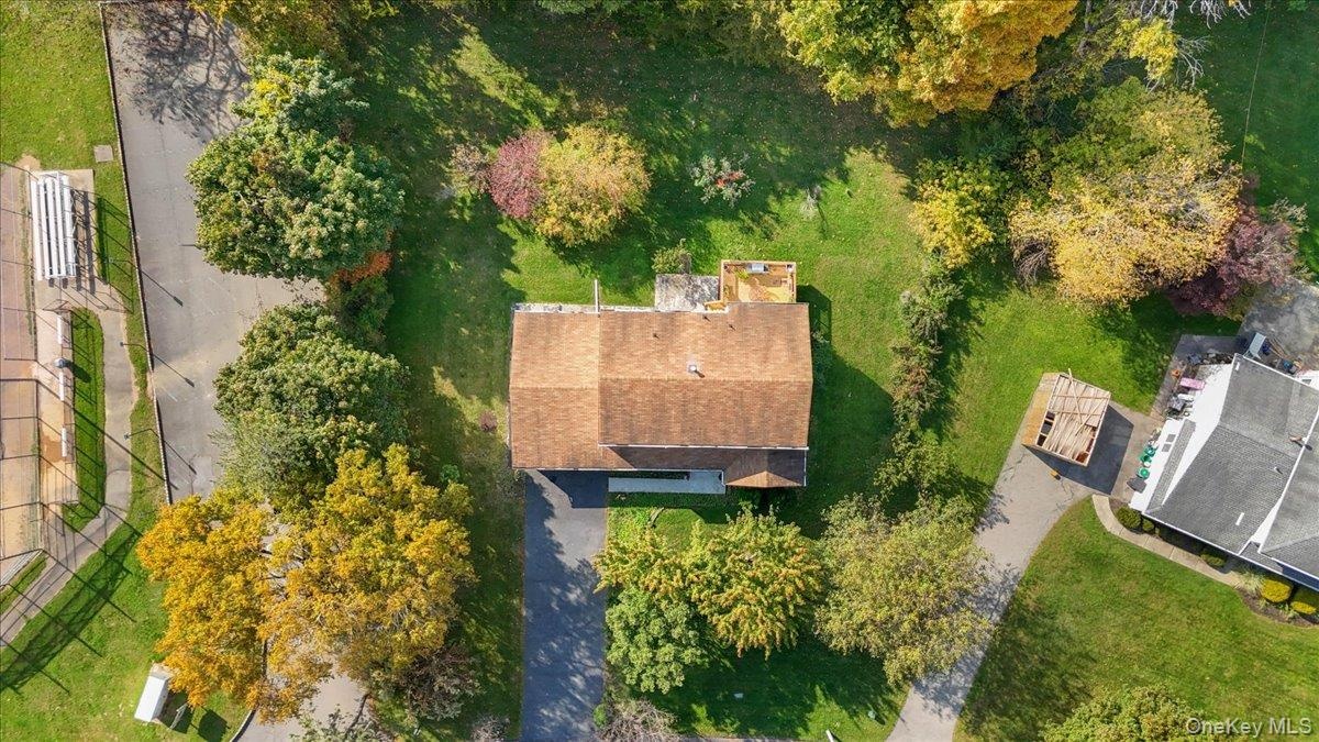 Aerial view of a residential lot featuring a shingled roof, paved driveway, expansive lawn, and mature trees