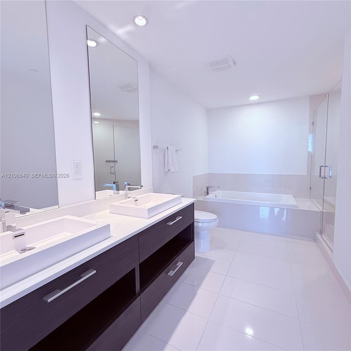 Dual vanity with rectangular vessel sinks and stone-composite countertop, dark wood-finish cabinetry, recessed ceiling lighting, tiled soaking tub, an