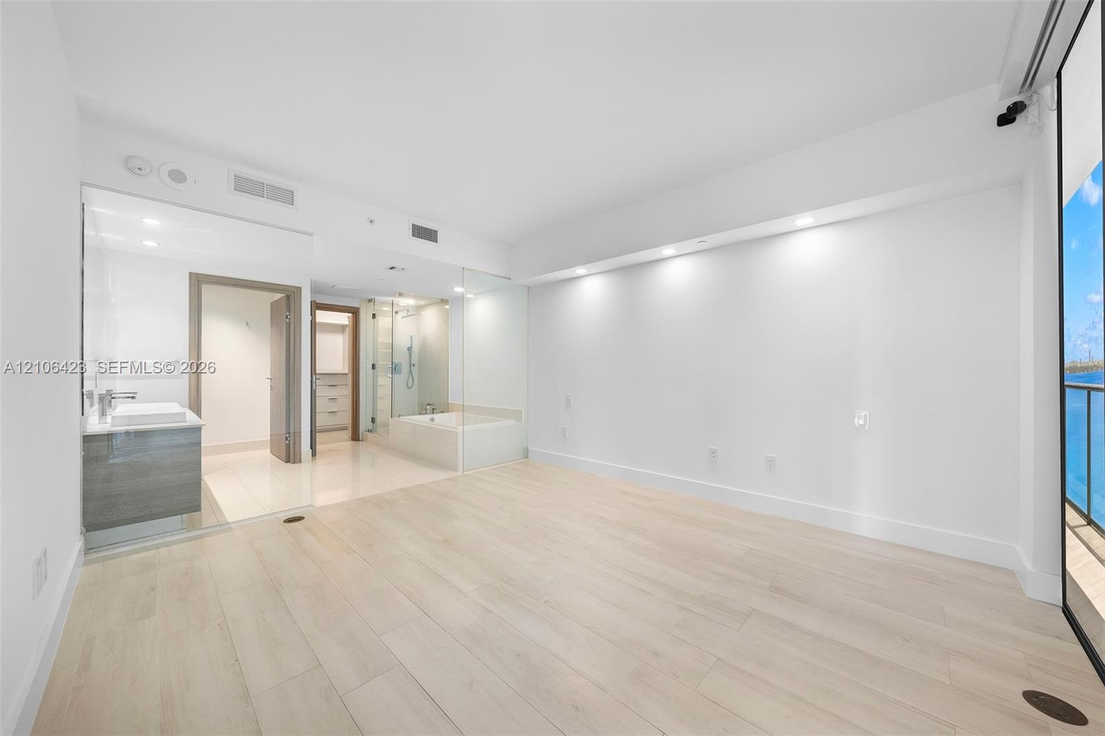 Open-concept space featuring light-toned wood-finish flooring, recessed architectural soffit lighting, glass-enclosed shower, soaking tub, and floor-t