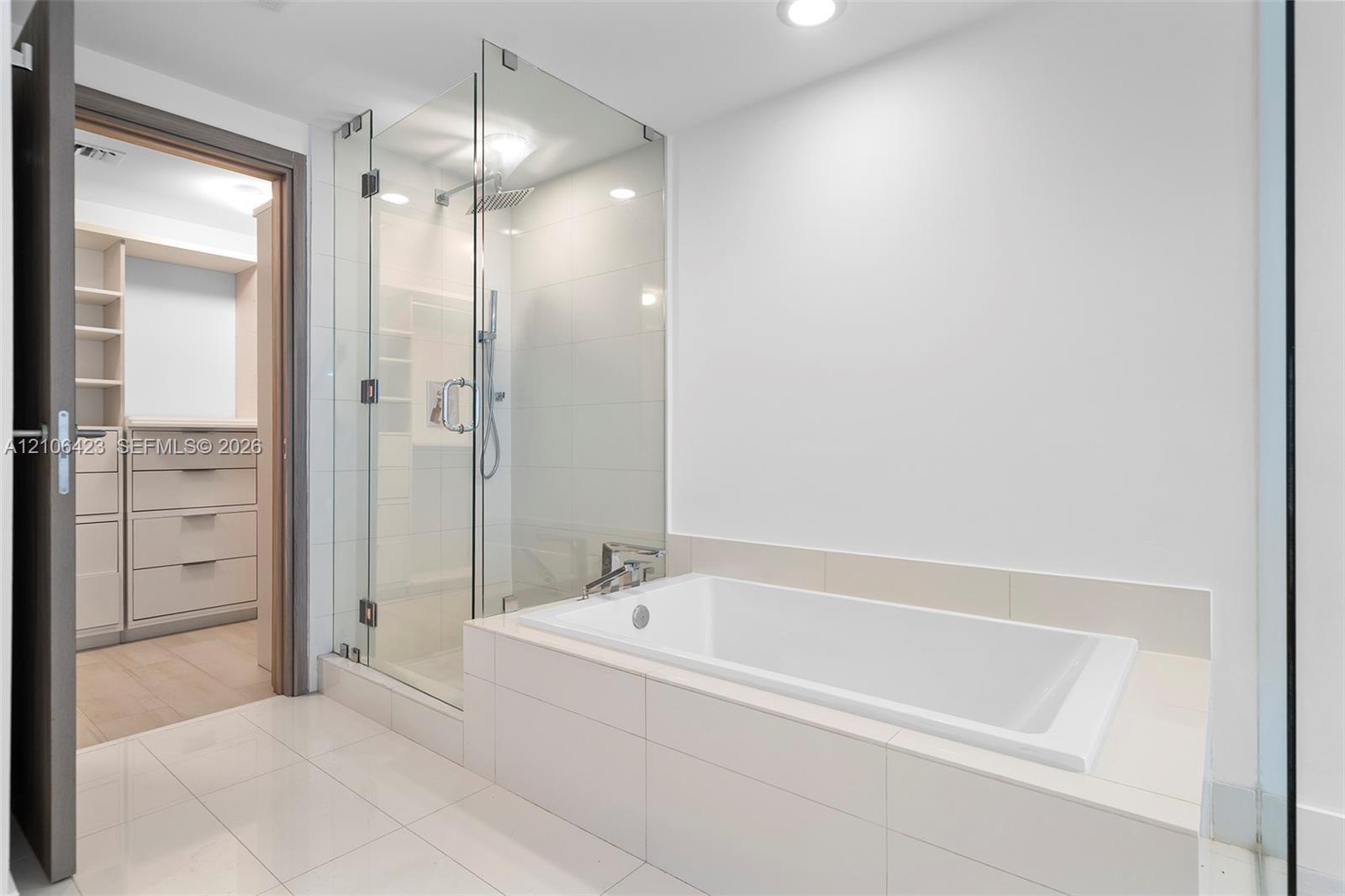 Glass-enclosed walk-in shower with rain showerhead, deep-soaking bathtub with tile surround, built-in custom closet cabinetry, and large-format floor 