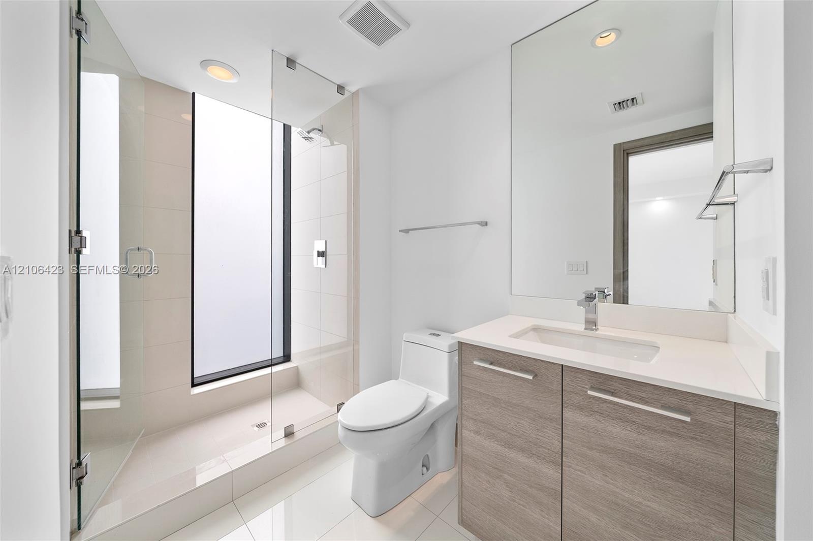 Full-height frameless glass shower enclosure with integrated window, wood-finish vanity with stone countertop, rectangular undermount sink, and polish
