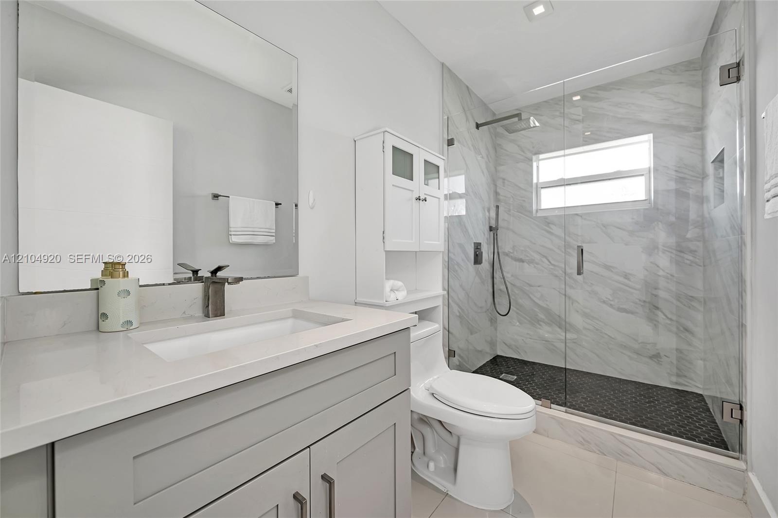 Bathroom featuring a grey vanity with a rectangular sink, stone-finish countertop, walk-in shower with floor-to-ceiling tile, and a wall-mounted glass