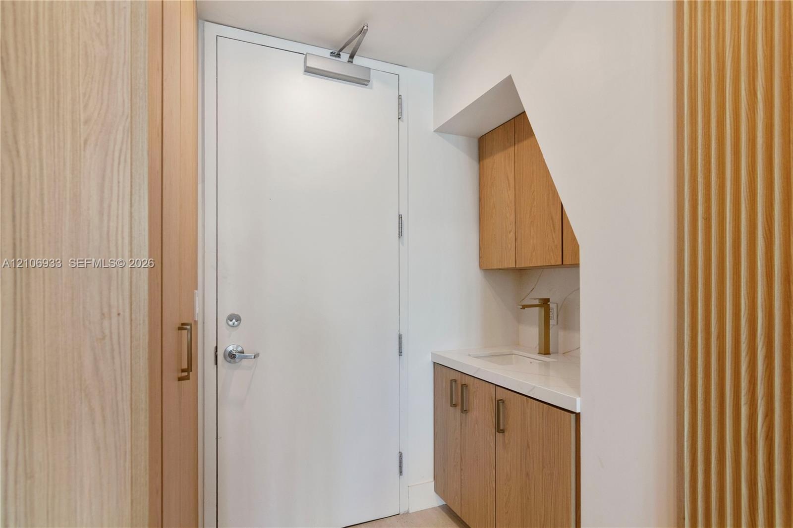 Built-in cabinetry featuring light wood-finish doors, stone-surface countertop, integrated sink, brushed gold-tone faucet, and floor-to-ceiling vertic