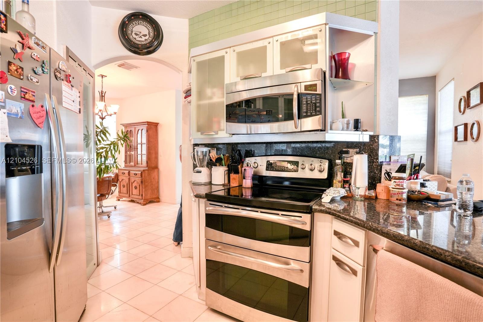 Kitchen featuring stainless steel appliances, dark stone-finish countertops, white cabinetry with glass-front uppers, and light-toned tile flooring