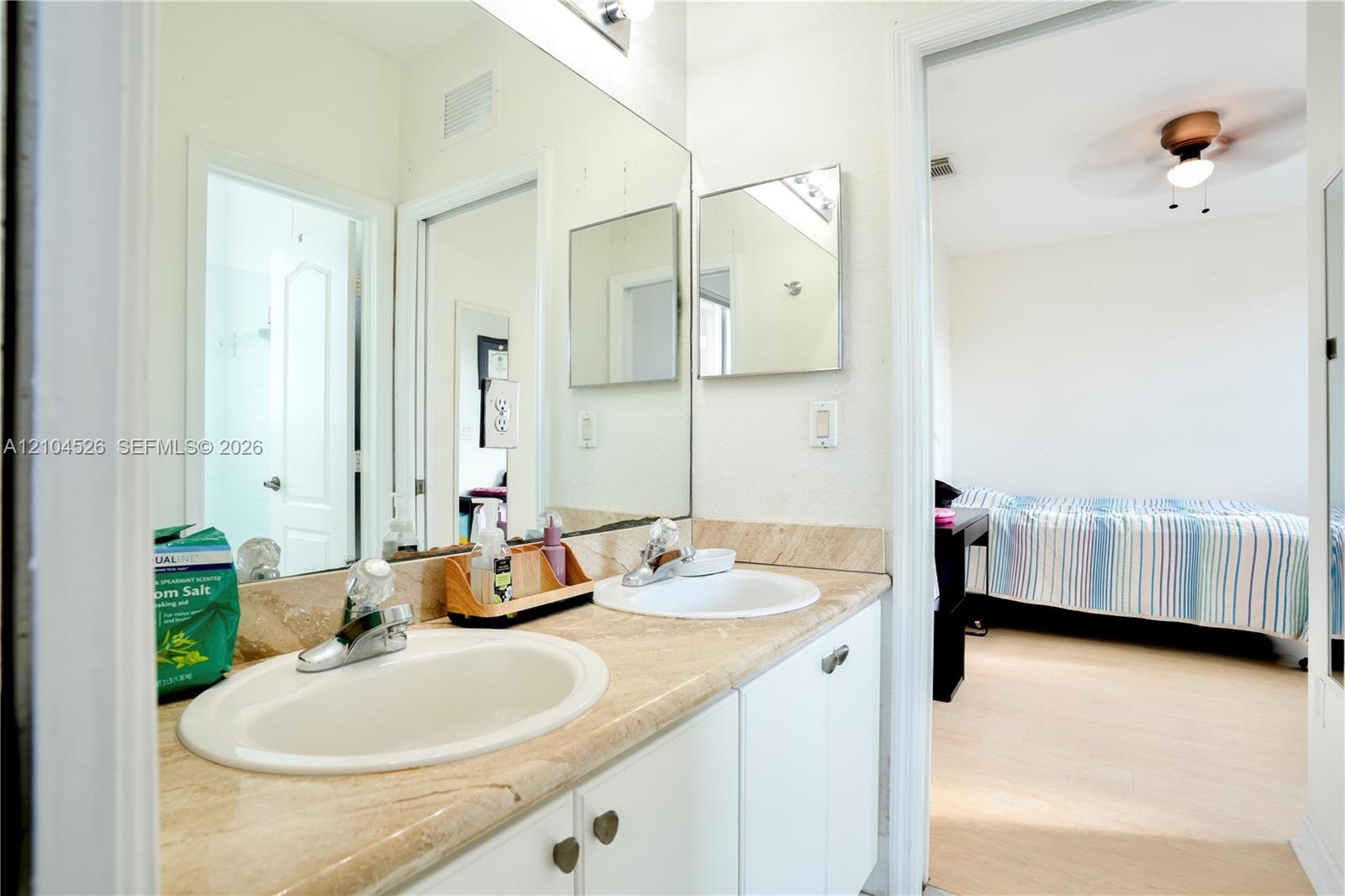 Dual-sink vanity with a natural stone countertop, white cabinetry, chrome-finish faucets, and an expansive wall-mounted mirror