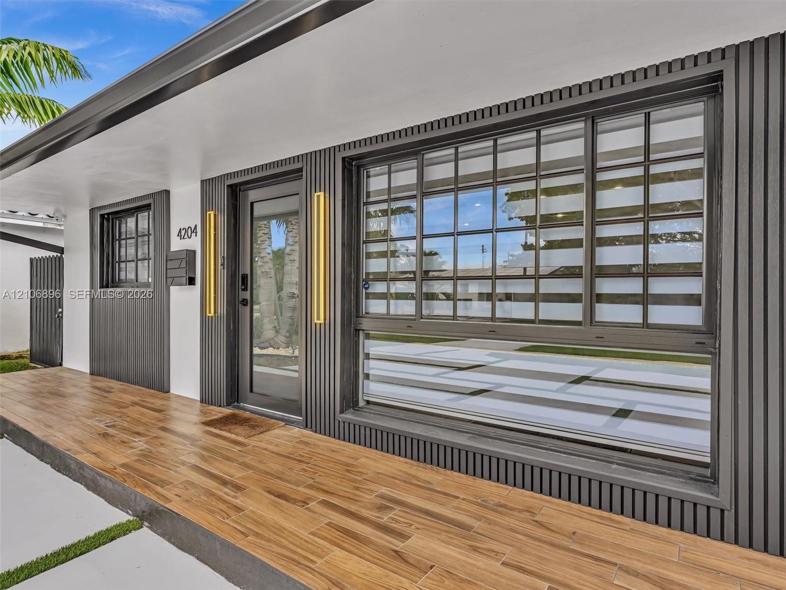 Exterior entry featuring wood-finish floor tiling, dark vertical slat siding, a glass-panel entry door, multi-pane sliding window, and dual vertical w