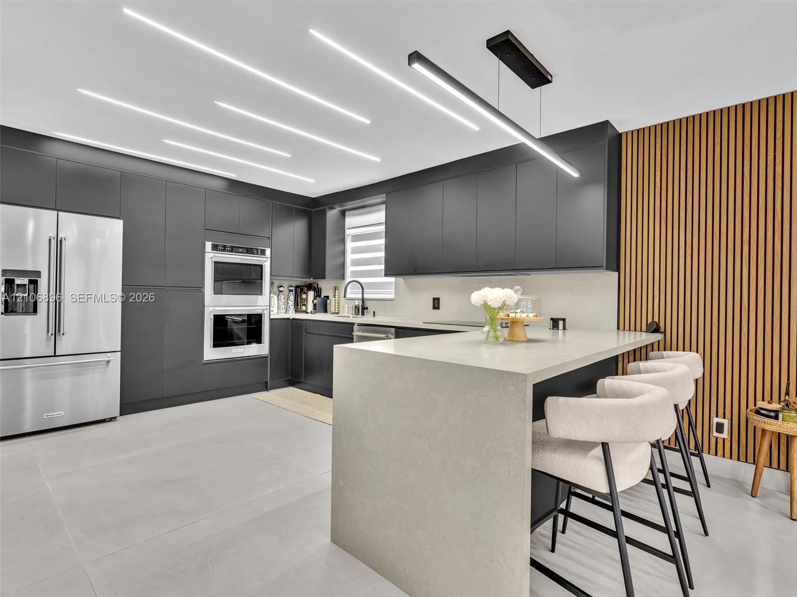 Modern kitchen featuring flat-panel cabinetry, a waterfall-edge stone island, linear LED ceiling lighting, integrated double wall ovens, and a vertica