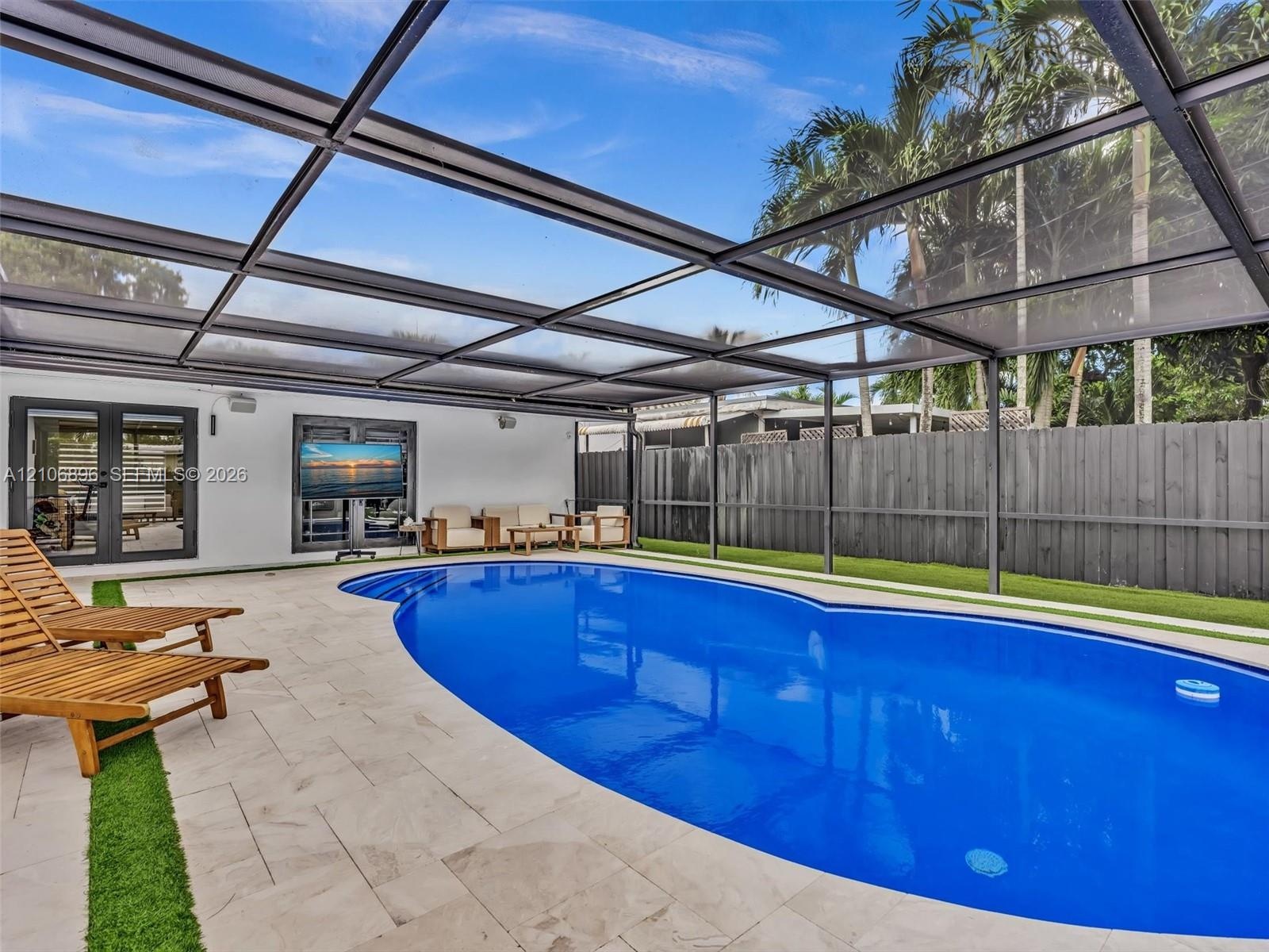 Screened pool enclosure featuring a custom-shaped swimming pool, light-toned stone pool deck with integrated turf detailing, and a high-privacy wood f