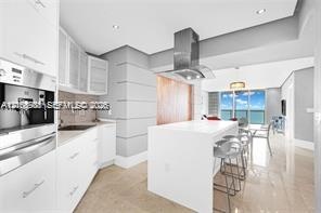 Modern kitchen featuring white flat-panel cabinetry, a central island with a solid-surface countertop, stainless steel vent hood, built-in coffee syst