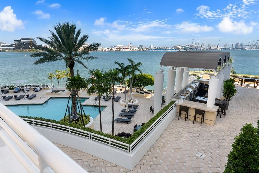 Waterfront pool terrace featuring a paved sun deck, built-in outdoor bar with a covered structure, mature palm trees, and perimeter landscape hedging