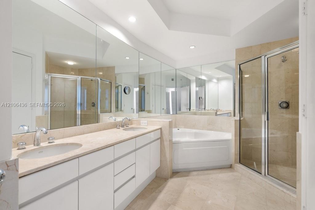 Double vanity with light-toned stone countertop, expansive wall-to-wall mirrors, recessed lighting, corner soaking tub, and enclosed shower with stone