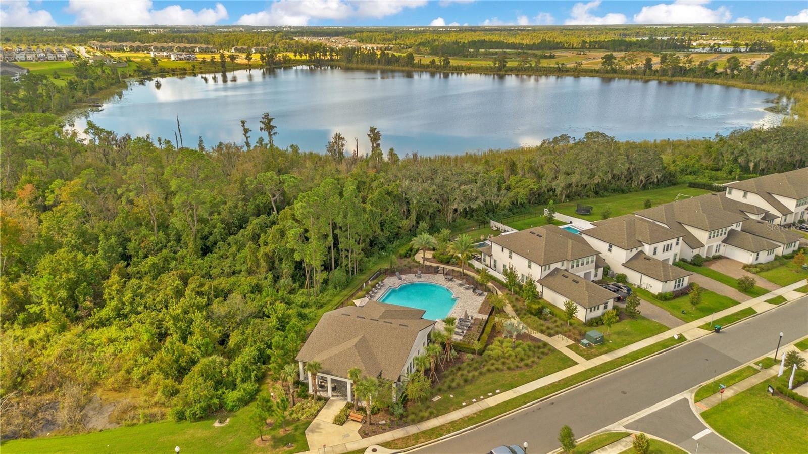 Aerial view of the community amenity area with a pool, clubhouse and lake backdrop.