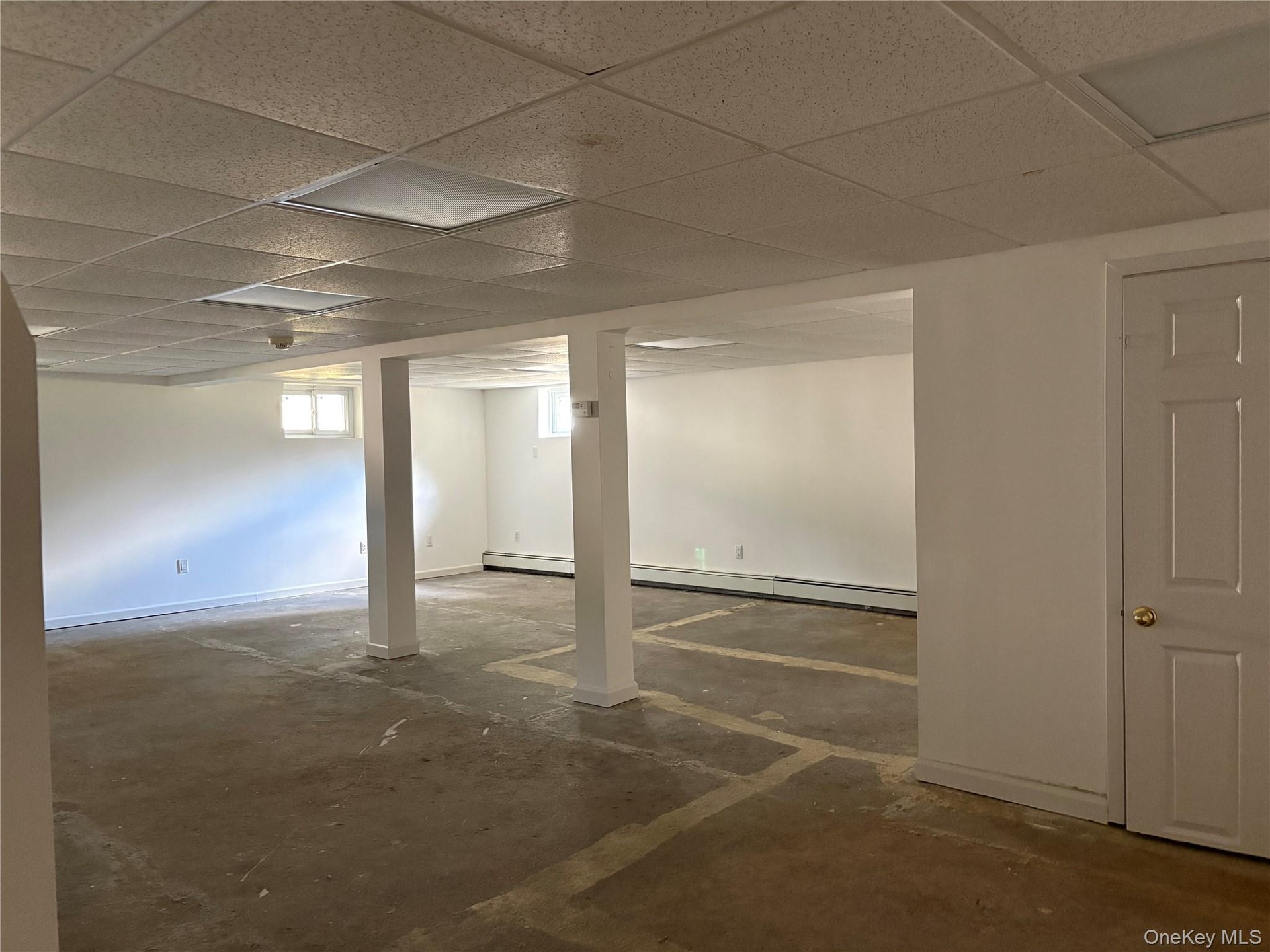 Open-concept partially finished basement space featuring white walls, drop ceiling panels, structural support columns, recessed windows, and closed of