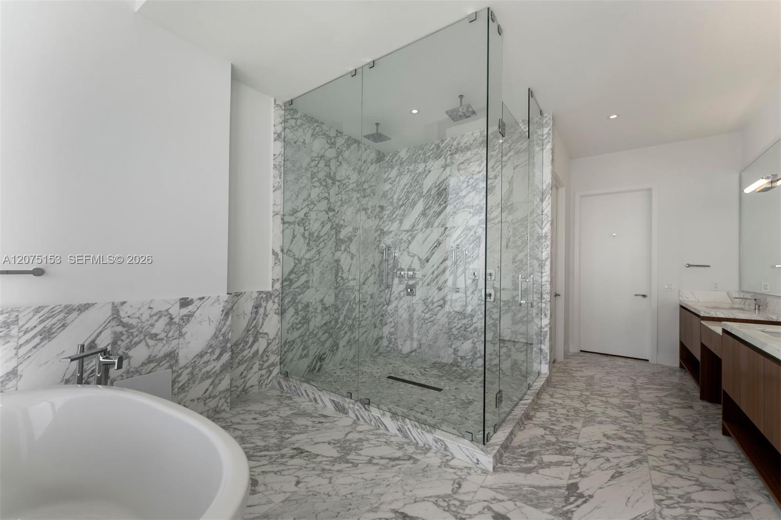 Walk-in glass-enclosed shower featuring floor-to-ceiling stone tile, freestanding soaking tub, dual-sink vanity with wood-finish cabinetry, and stone 