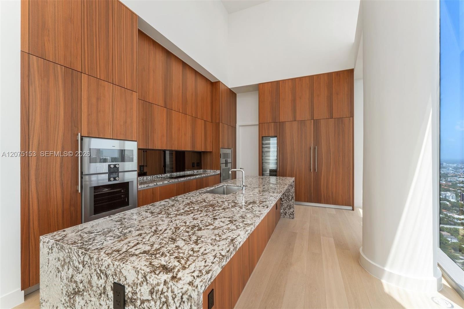 Kitchen featuring full-height wood-finish cabinetry, stone countertop island with integrated stainless steel sink, built-in stainless steel double wal