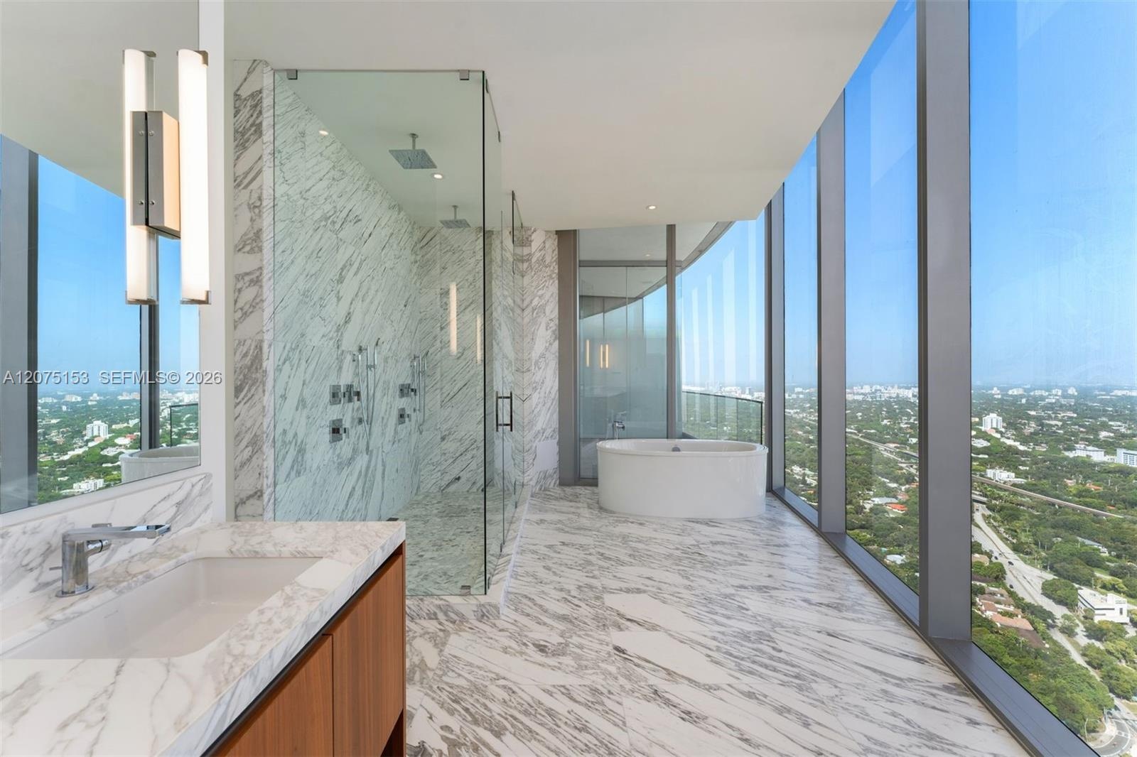 Floor-to-ceiling windows, natural stone surfaces, walk-in glass-enclosed shower with dual rainfall showerheads, and freestanding soaking tub