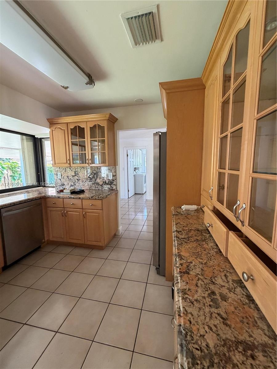 Kitchen featuring light-finish cabinetry with glass-panel upper doors, stone-surface countertops, stone-pattern backsplash, stainless steel dishwasher