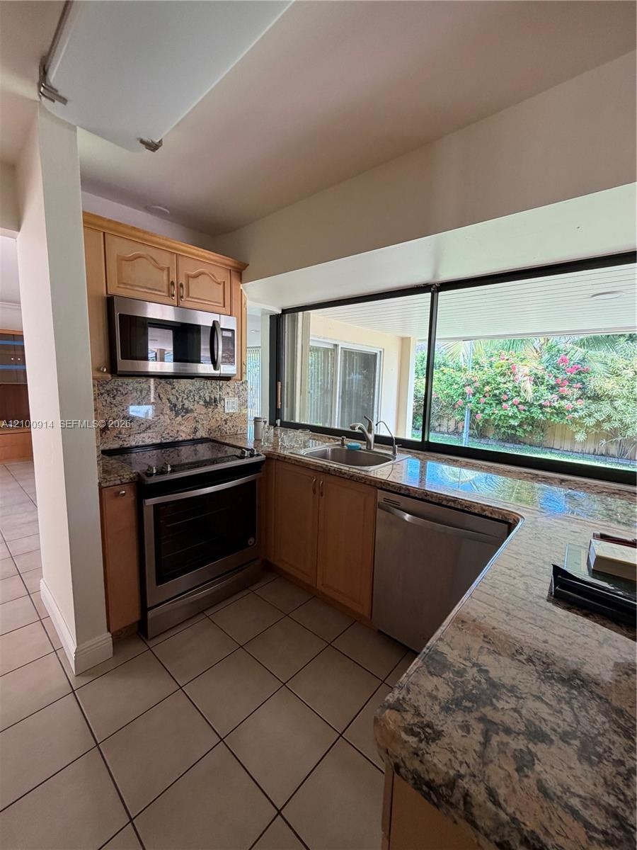 Kitchen featuring natural wood cabinetry, stone countertops, stainless steel appliances, tile flooring, and a large window