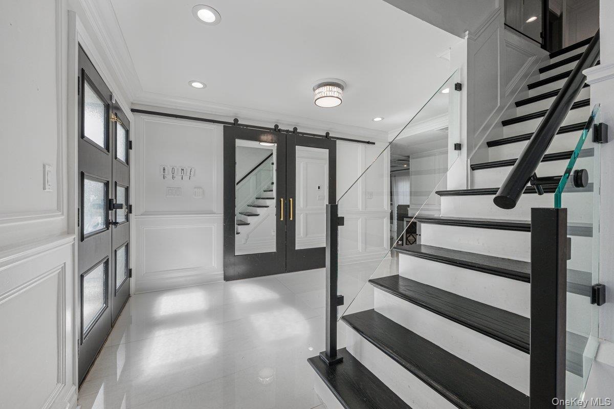 Foyer featuring high-gloss tile flooring, wainscoting, glass-panel entry door, sliding barn-style door with inset mirror, and staircase with glass rai
