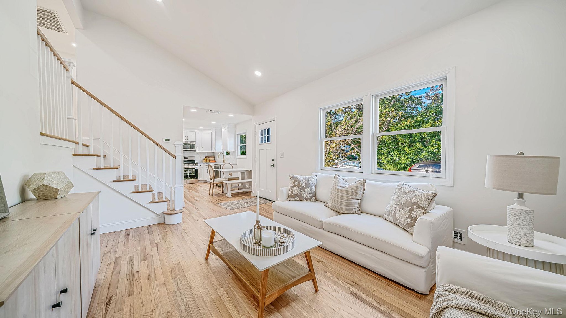 Living room with vaulted ceilings, wood-finish flooring, staircase with white railings, and large windows providing natural light
