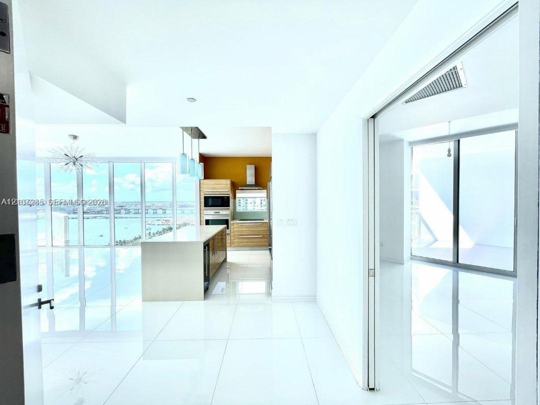 Open-concept kitchen featuring light wood-finish cabinetry, a central island with a stone countertop, built-in stainless steel appliances, white tile 