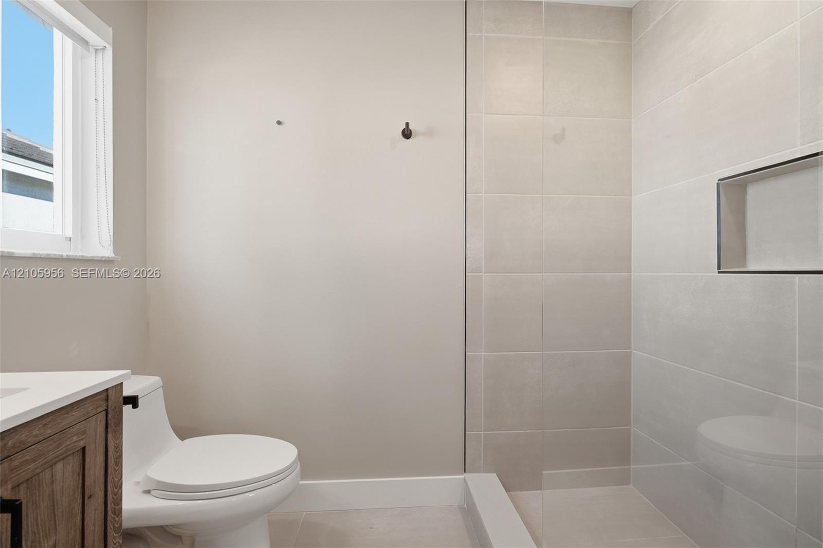 Wood-finish vanity, white countertop, walk-in shower with large-format wall tile, glass partition, and recessed shower niche