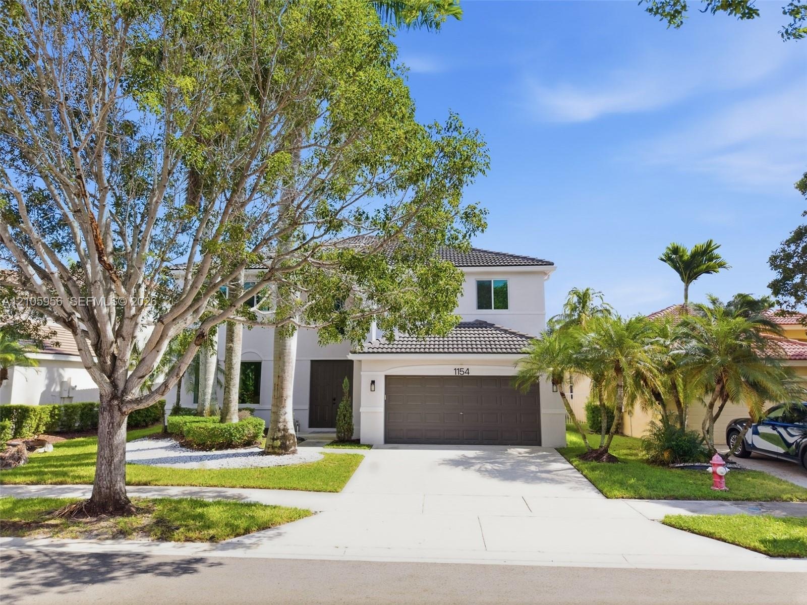 Two-story exterior featuring a light-colored stucco facade, tile roofing, an attached two-car garage, and established landscaping with mature trees an