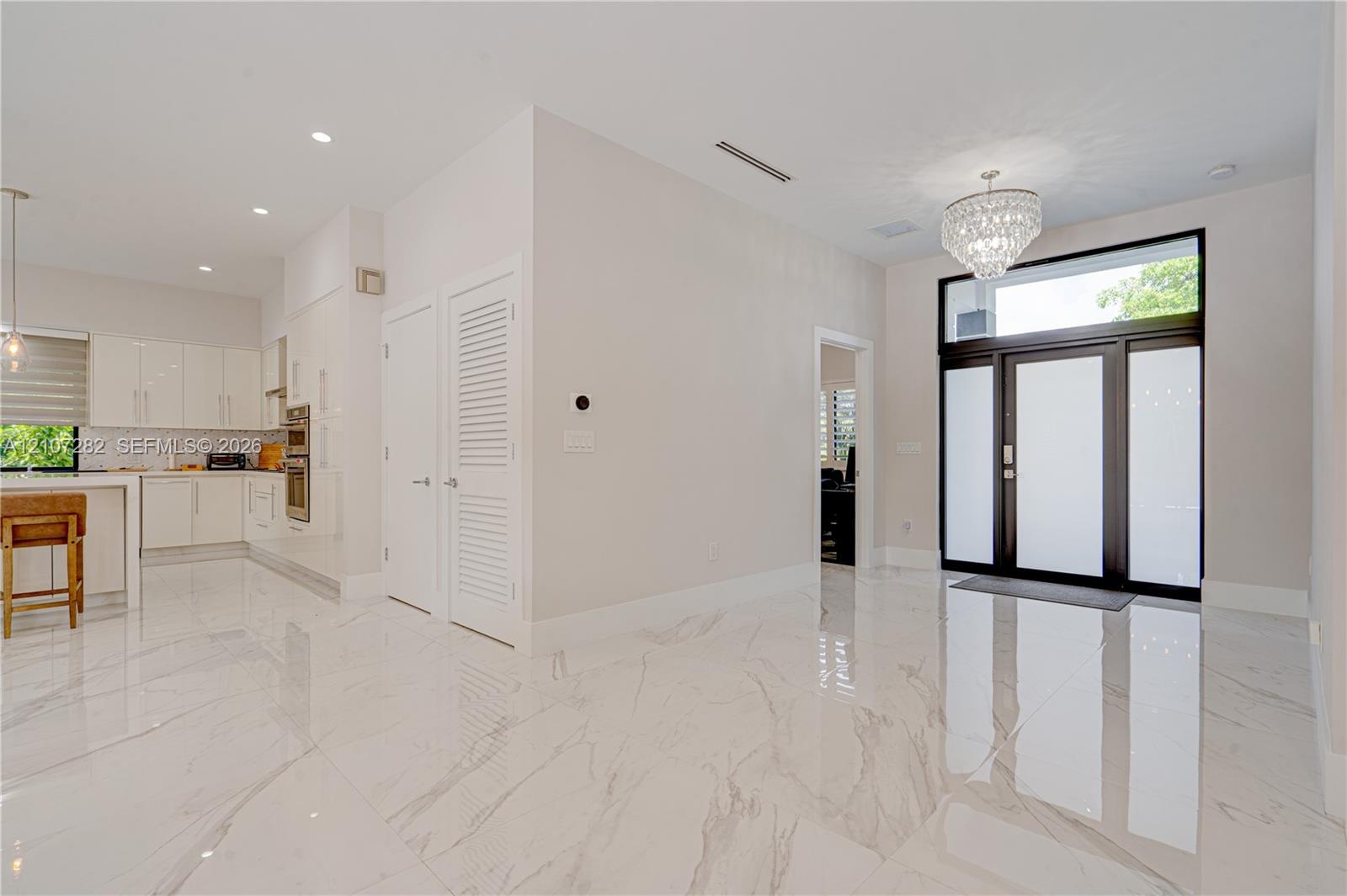 Large-format polished tile flooring, glass-panel front door with transom window, high-gloss white cabinetry, integrated wall oven, and tiered crystal 