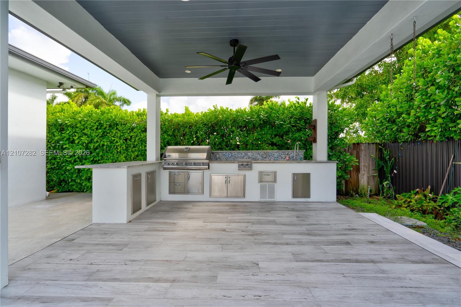 Covered outdoor kitchen featuring wood-finish tile flooring, built-in stainless steel grill, stone-style countertop, mosaic tile backsplash, and overh