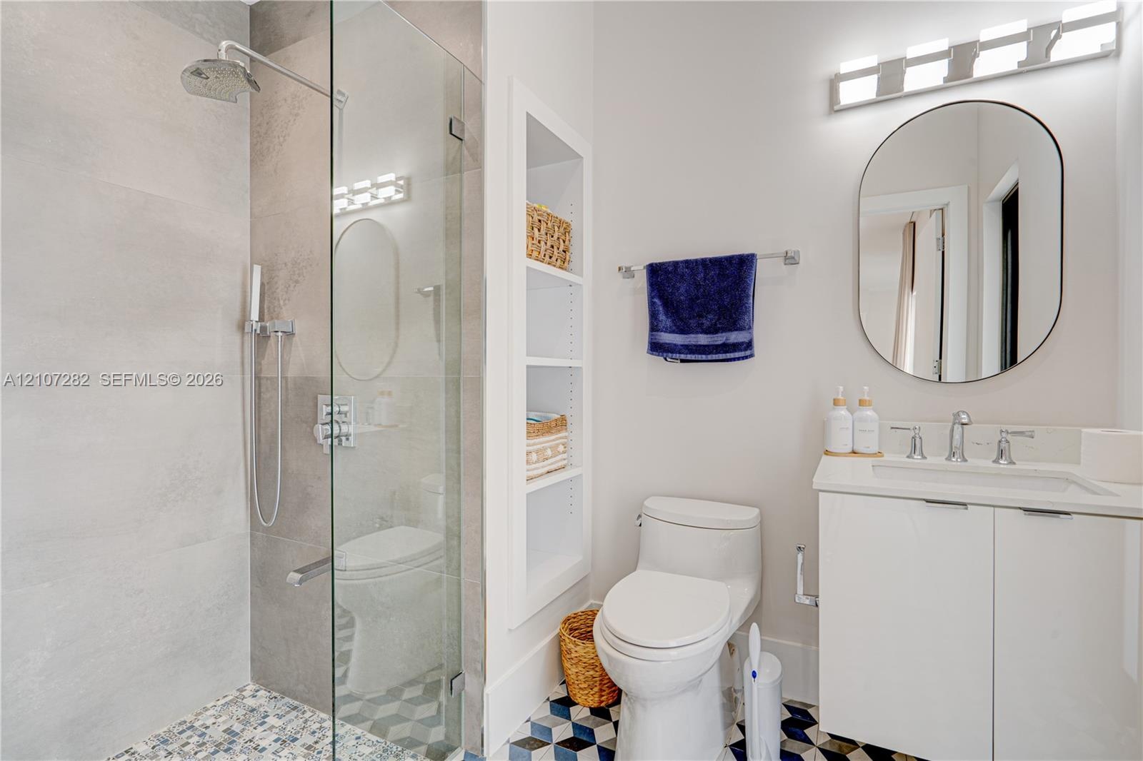 Bathroom featuring a glass-enclosed shower with a rainfall showerhead, geometric patterned floor tile, integrated wall shelving, an oval wall-mounted 