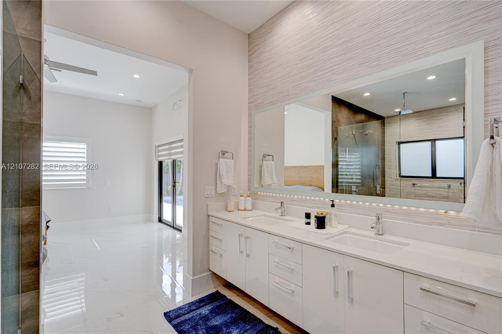 Dual-sink vanity with white cabinetry, light stone-finish countertops, full-width mirror with integrated lighting, and high-gloss tile flooring
