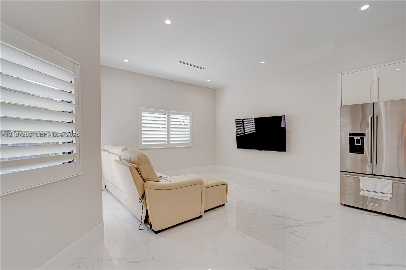 Open-concept area featuring white tile flooring, recessed ceiling lighting, plantation shutters, and a built-in stainless steel refrigerator with over