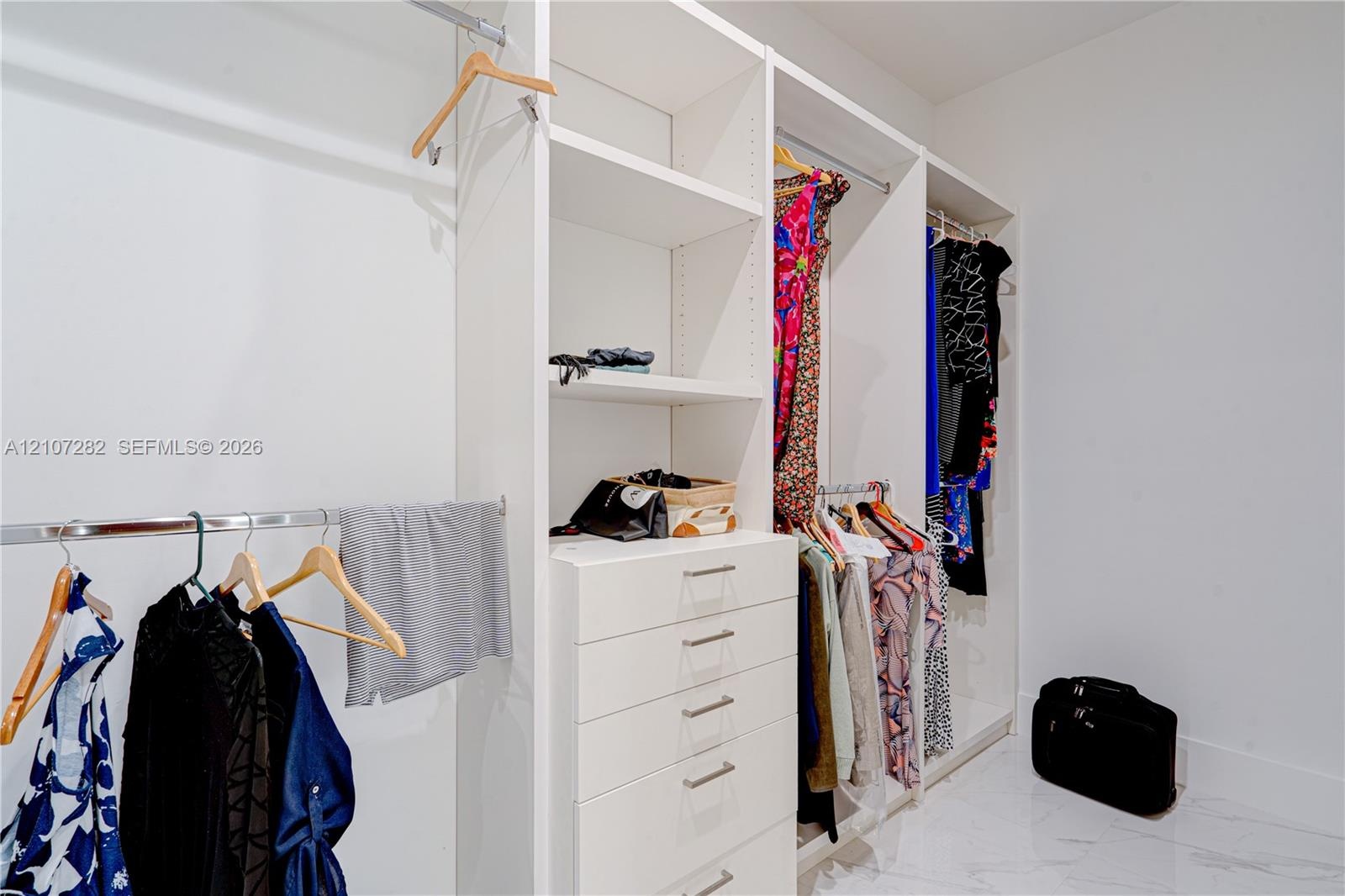 Custom walk-in closet featuring white built-in shelving, a central column of five soft-close drawers, stainless steel hanging rods, and polished stone