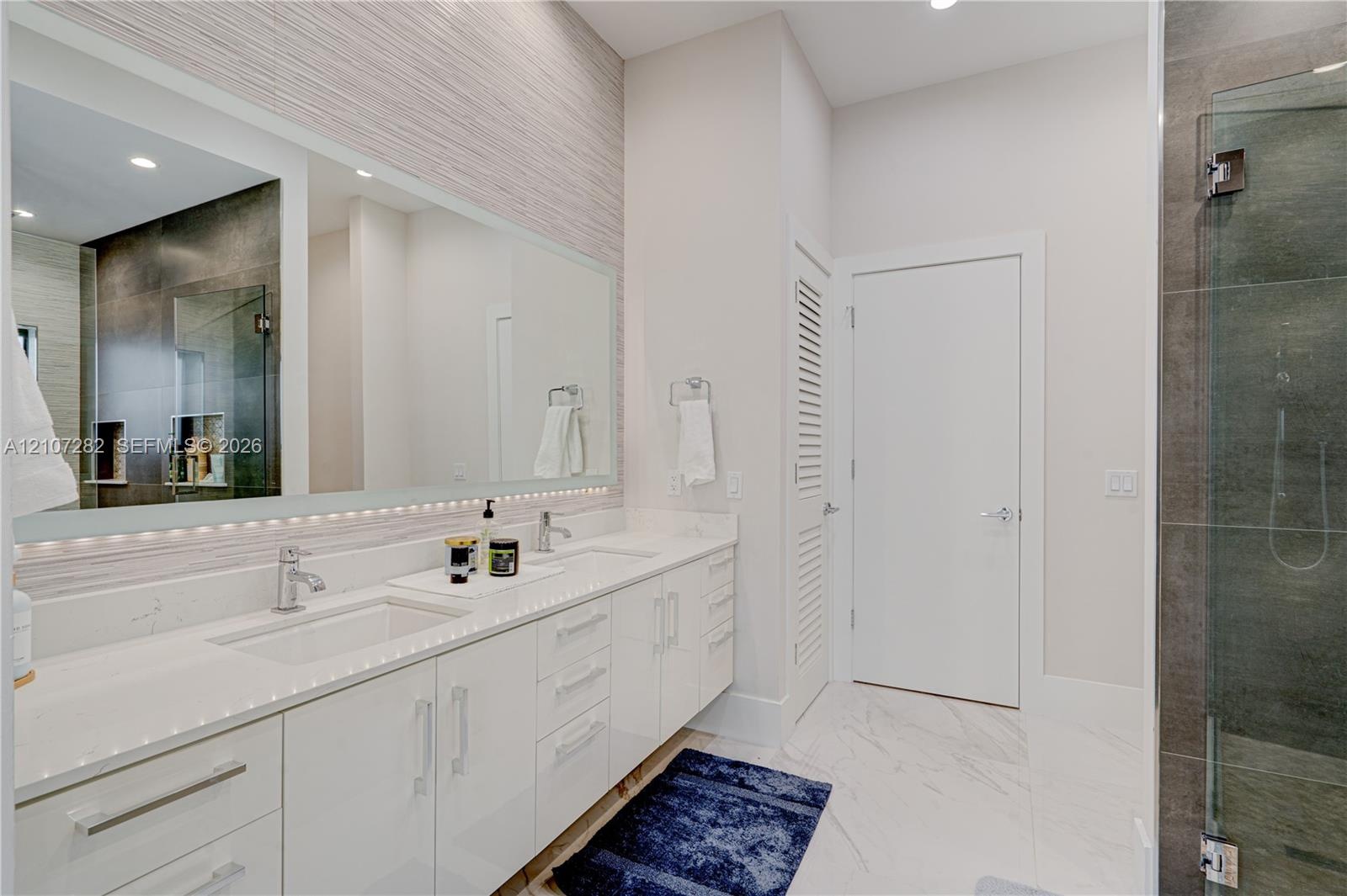 Double vanity with white cabinetry, light-toned stone-finish countertops, undermount sinks, large wall-spanning mirror, and a walk-in glass-enclosed s