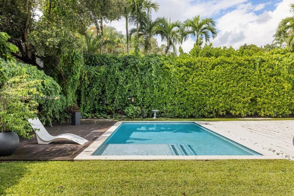 Rectangular swimming pool featuring a light-toned stone surround, wood-finish decking, and an expansive backdrop of manicured hedges and mature palm t