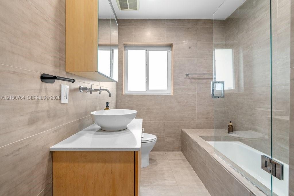 Bathroom featuring floor-to-ceiling neutral tile, custom wood-finish vanity with a white vessel sink, quartz countertop, wall-mounted faucet, and a gl