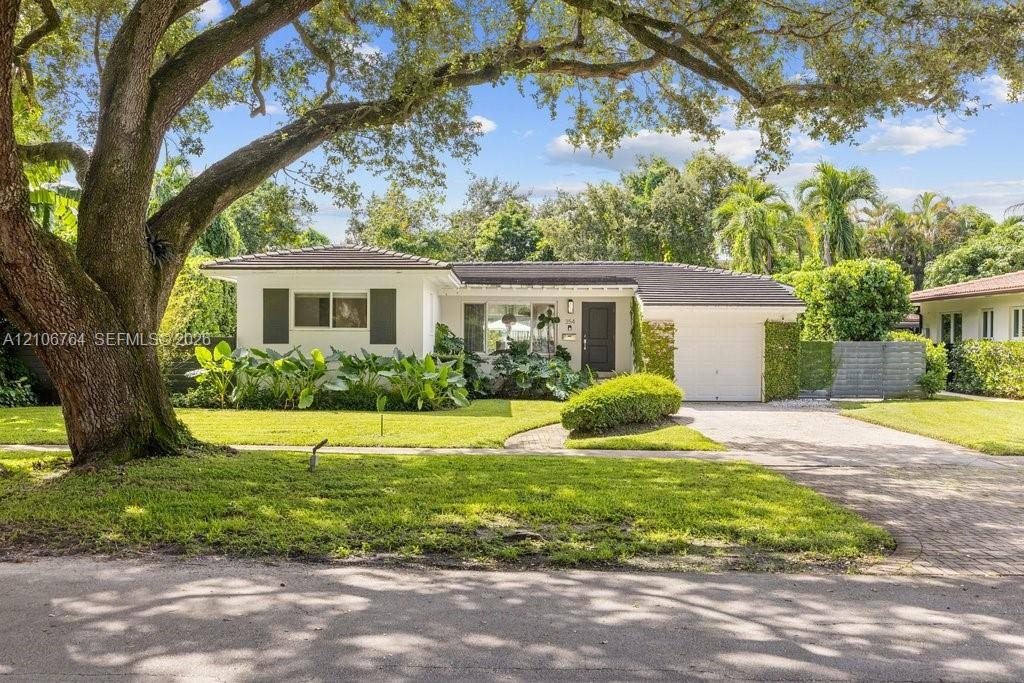 Single-story exterior featuring a light-toned stucco façade, dark-toned roof shingles, a paved driveway, and established landscape elements including 