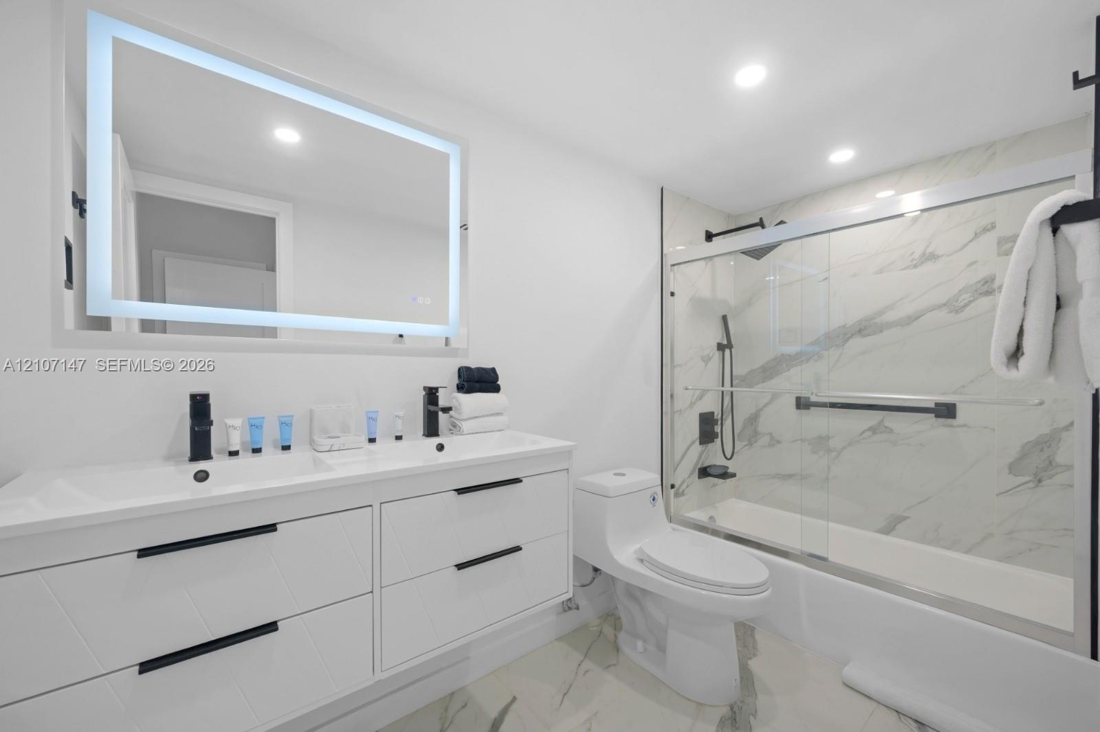 Modern bathroom featuring a double-sink vanity with matte black hardware, backlit LED mirror, recessed ceiling lighting, and a bathtub-shower combinat