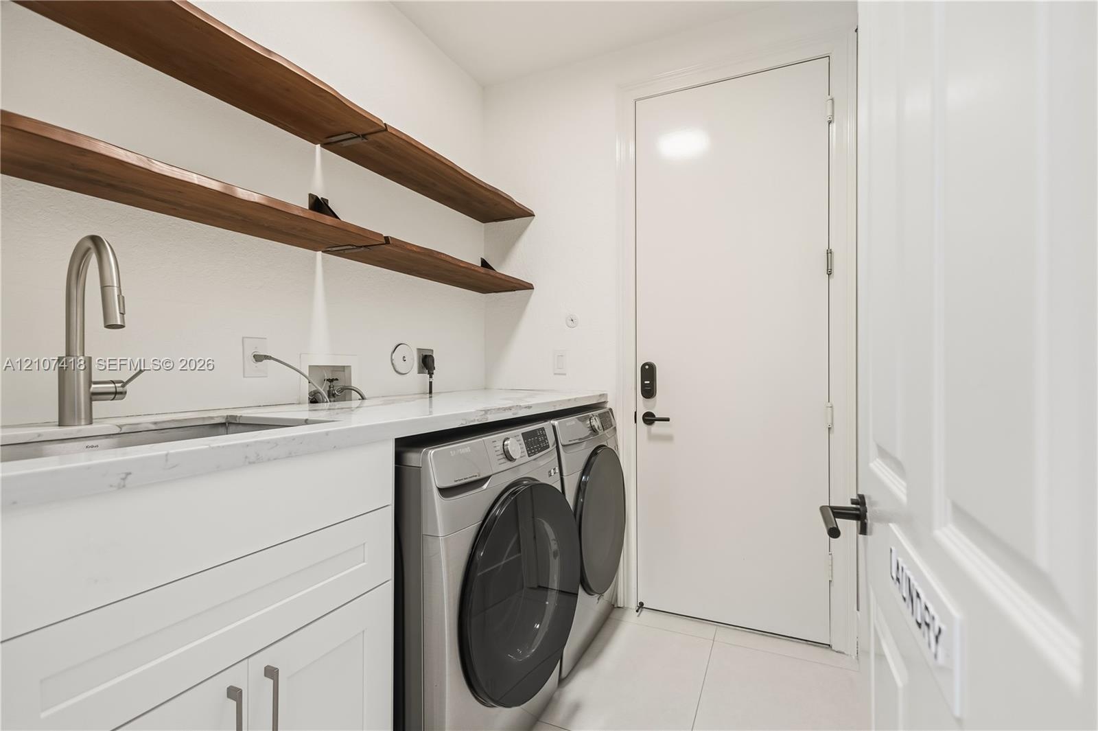 Laundry room featuring white cabinetry, stone-finish countertops, an undermount sink with a high-arch faucet, wall-mounted wood-finish shelving, and l