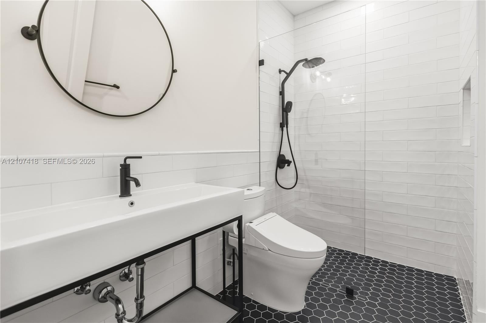 Wall-mounted round mirror, console-style vanity with a rectangular sink, matte black faucet hardware, glass-enclosed shower with subway tile surround,