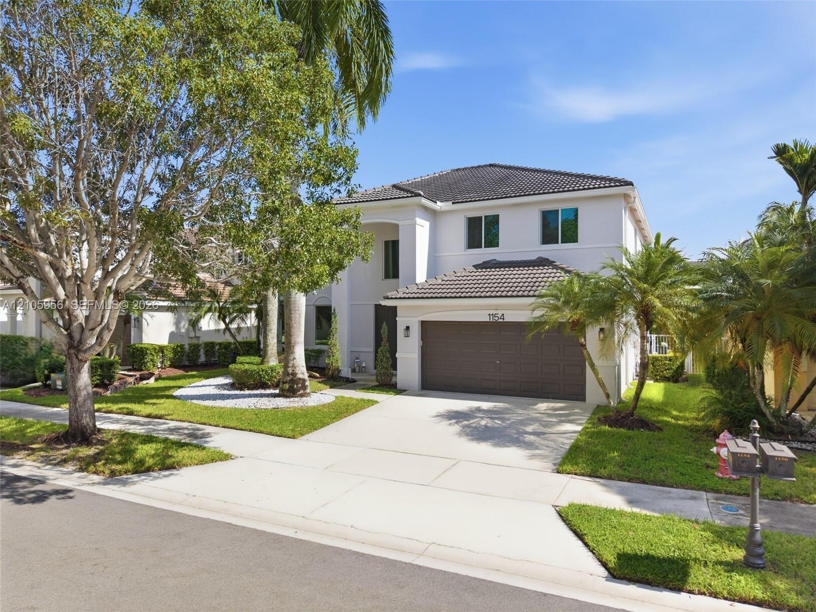 Two-story stucco exterior featuring a concrete tile roof, attached two-car garage, and mature professional landscaping