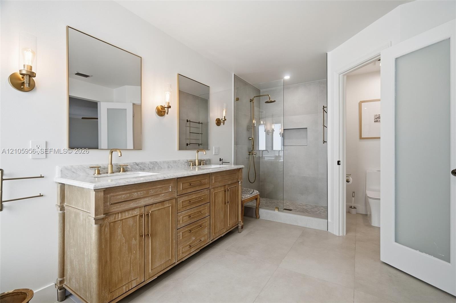 Dual-sink vanity with stone countertop, walk-in glass-enclosed shower featuring gold-tone hardware, wall-mounted light fixtures, and frosted glass doo