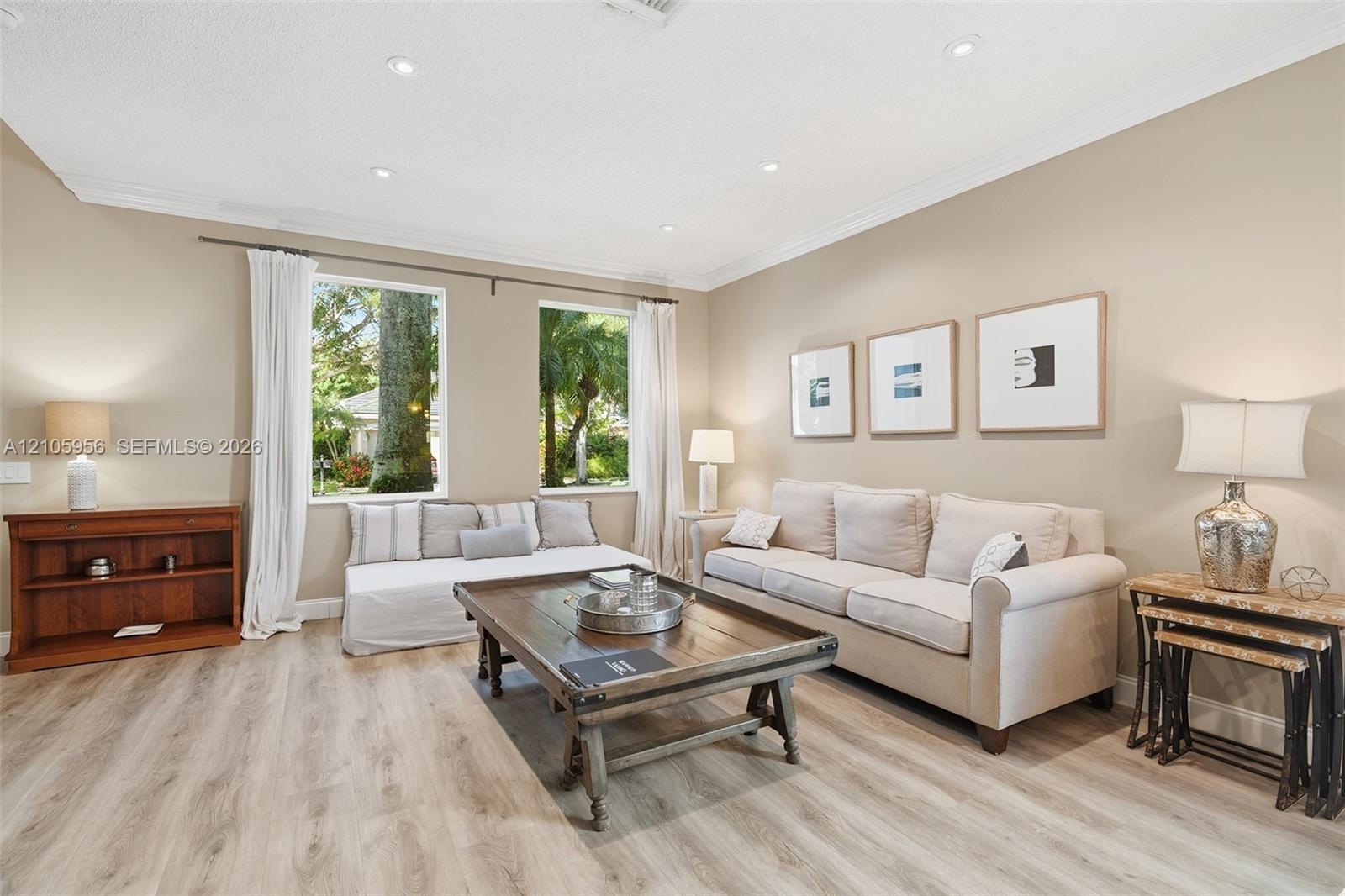 Living space featuring light-tone wood-finish flooring, recessed lighting, crown molding, and large rectangular windows with exterior greenery