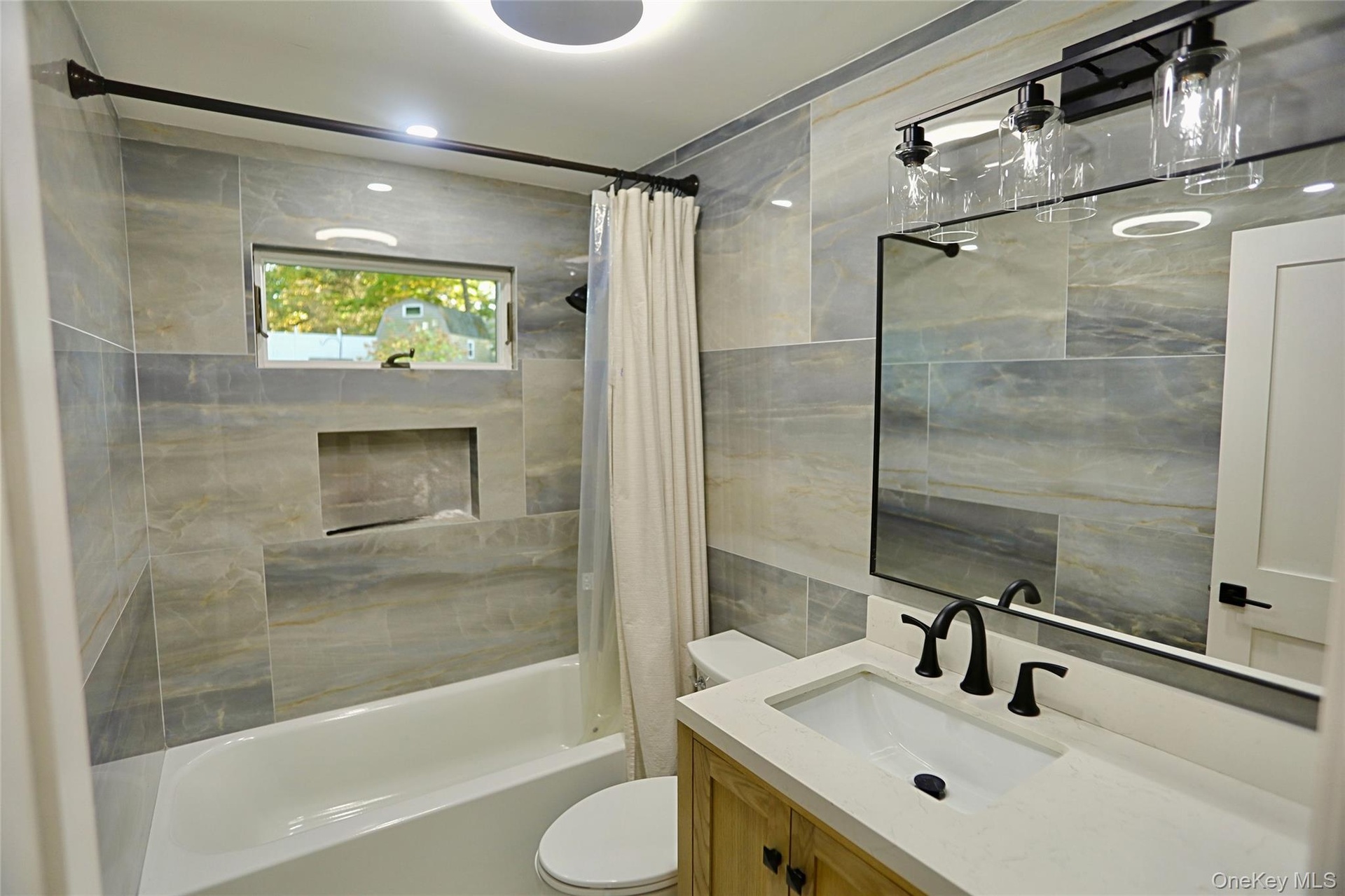 Bathroom featuring large-format neutral-toned wall tile, wood-finish vanity with light-colored solid surface countertop, integrated shower niche, and 